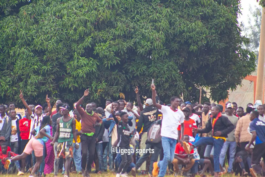 A crowd of Ligega Sports supporters celebrating with their arms raised under a large mango tree beside the pitch.