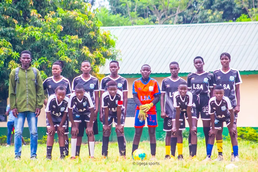 The Ligega Sports girls squad in black and white kit, lined up in two rows on the pitch with a coach standing at the end of the row.