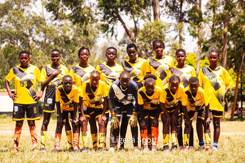 The Ligega Sports girls squad in yellow and black kit, lined up in two rows on a grass pitch under trees.