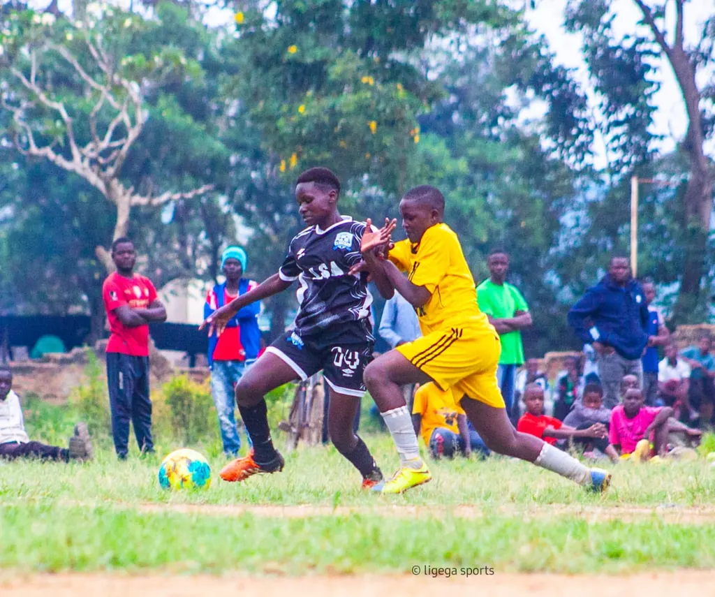 A Ligega Sports player in black kit shielding the ball from an opponent in yellow during a match, with spectators watching from the touchline.