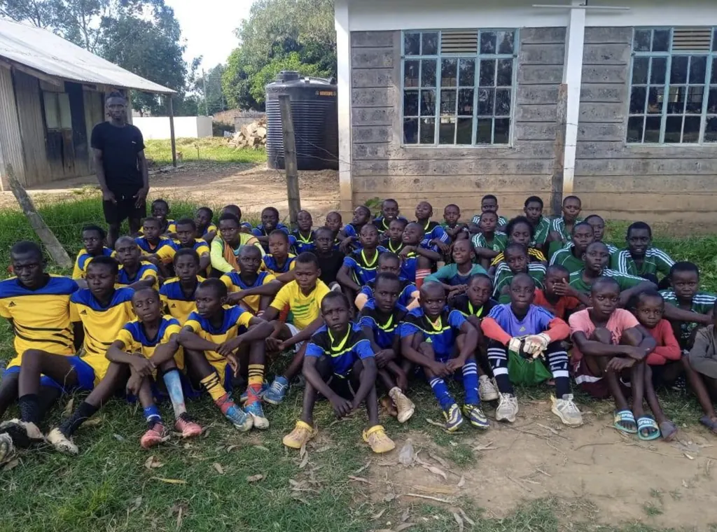 A large group of Ligega Sports juniors sitting together on the grass in training kit, with a coach standing behind them.