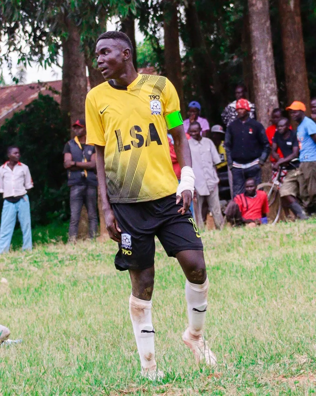 Stephen Ochieng Omondi wearing a green captain's armband, walking across the pitch in Ligega Sports yellow and black kit during a match.