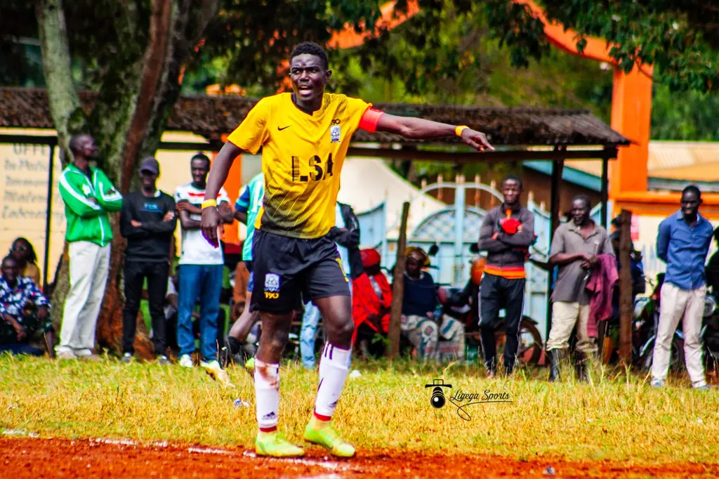 Stephen Ochieng Omondi in Ligega Sports yellow and black kit, arm raised giving instruction during a match, with spectators lining the touchline.