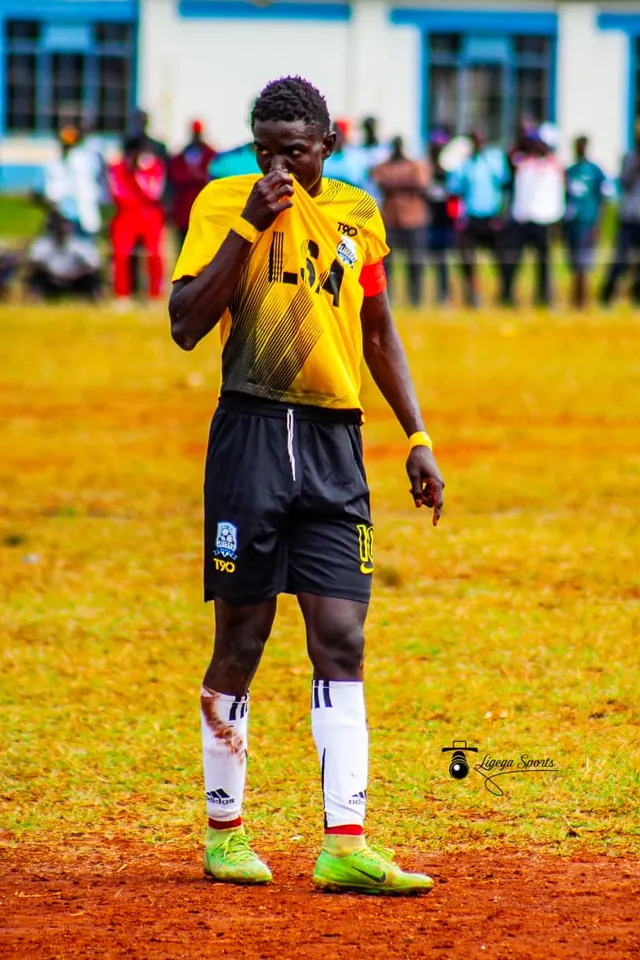 Stephen Ochieng Omondi kissing his Ligega Sports shirt on the pitch, with spectators watching in the background.