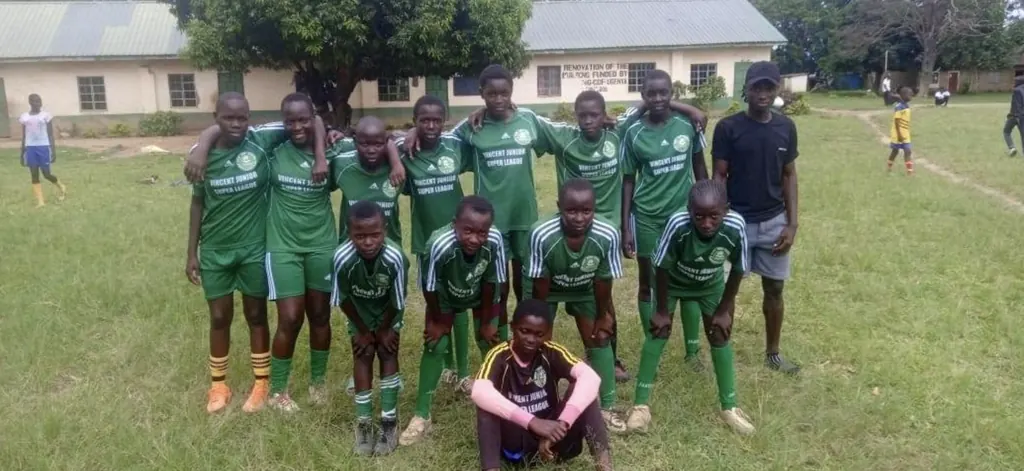 The Ligega Sports Starlets squad in green kit, photographed together on the grass outside a school building.