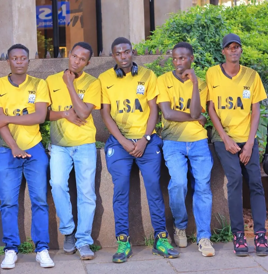 Five young men in yellow Ligega Sports LSA shirts standing against a wall in town.
